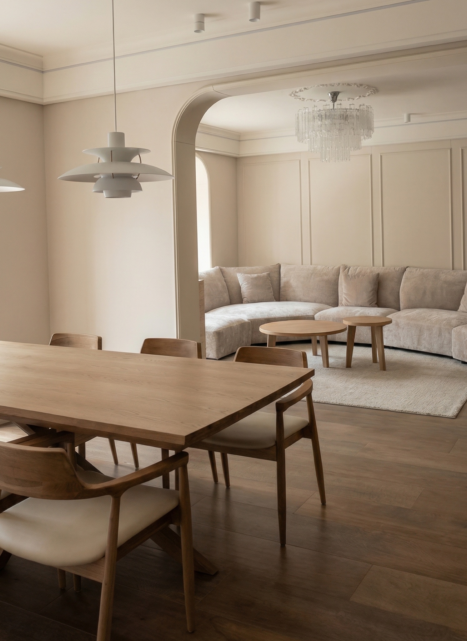 Living room with a cream curved sofa, round oak coffee tables and a crystal chandelier, seen through an arched opening from the dining room